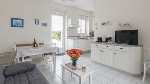 A modern, well-lit living room with a grey sofa, a dining table with chairs, a kitchen area with white cabinets, a TV, and a small table with a vase of flowers. - La Birochère - 400 m plage - Parking (Pornic)