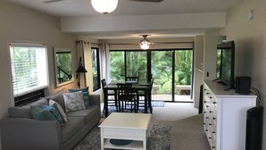A living room with a sofa, coffee table, and a dining area with a table and chairs. There is a ceiling fan, a television, and a window with blinds.