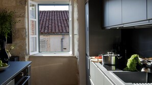 A modern kitchen with a stone wall, wooden ceiling beams, and a window with a red-tiled roof view.