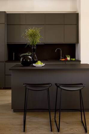 Une cuisine moderne avec un îlot central, deux tabourets de bar, un vase avec des plantes et un saladier rempli de fruits.