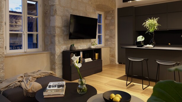 A modern living room with a stone wall, a flat-screen TV, a sofa, a coffee table with a book and a vase, and a kitchen area with stools.