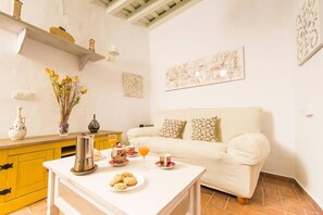 Een gezellige woonkamer met een witte bank, een bijzettafeltje met ontbijtartikelen, een geel kastje en decoratieve wanddecoratie.