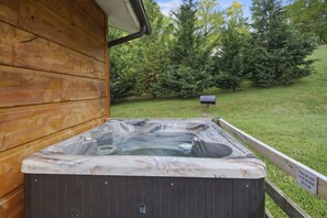 Un jacuzzi al aire libre frente a una cabaña de madera, con un jardín césped y árboles de fondo.