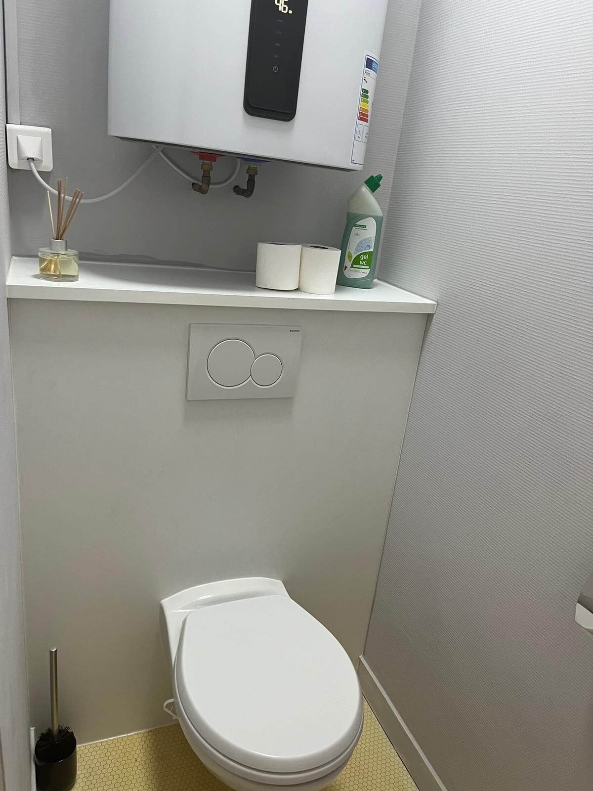 A small, modern bathroom with a toilet, a wall-mounted toilet paper holder, a hand soap dispenser, and a toilet brush.