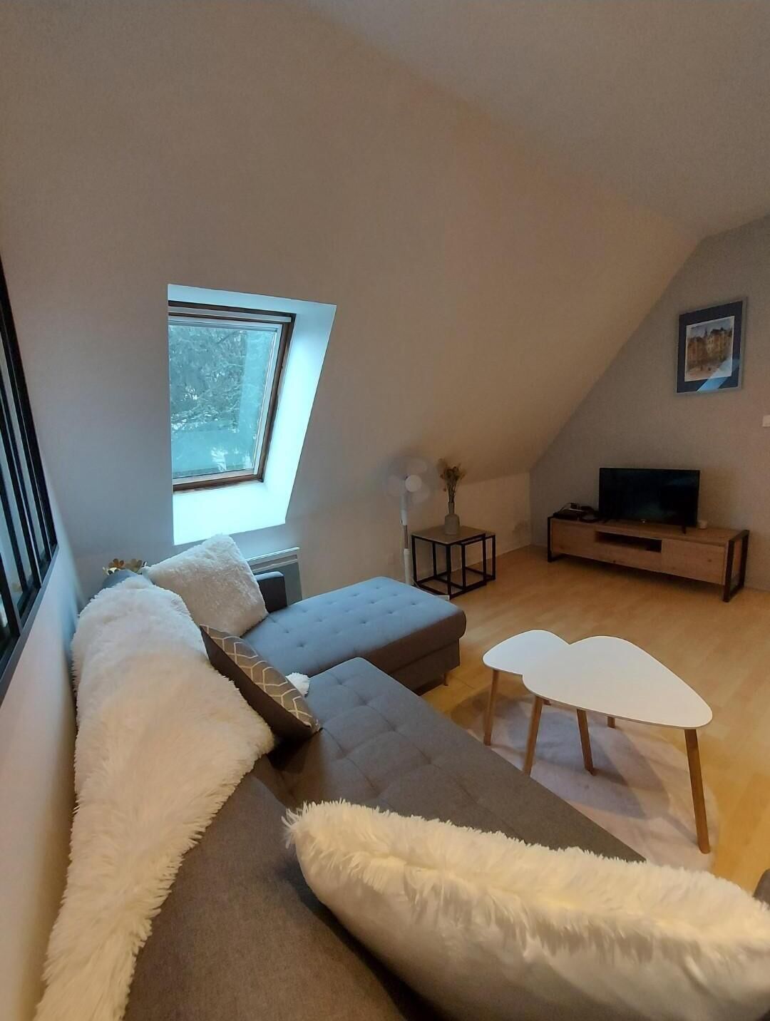 Ein modernes Wohnzimmer im Loft-Stil mit einem großen Fenster, einem grauen Ecksofa, einem weißen Couchtisch und einem Fernseher auf einem Holzgestell.