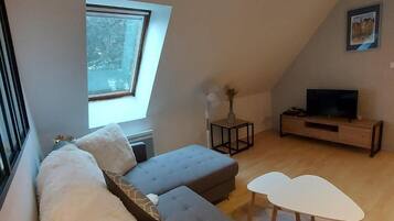 A modern loft-style living room with a large window, a grey sectional sofa, a white coffee table, and a TV on a wooden stand.