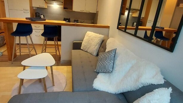 Una sala moderna con un sofá gris, dos almohadones blancos y mullidos, y una mesita auxiliar blanca. Se ve una cocina en el fondo con armarios blancos y una encimera de madera.