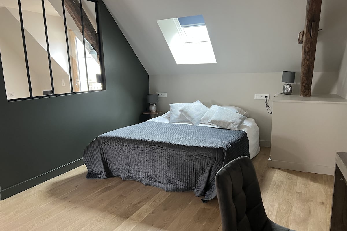 A bedroom with a sloped ceiling, a skylight, a bed with a grey quilt, a bedside table with a lamp, and a chair.