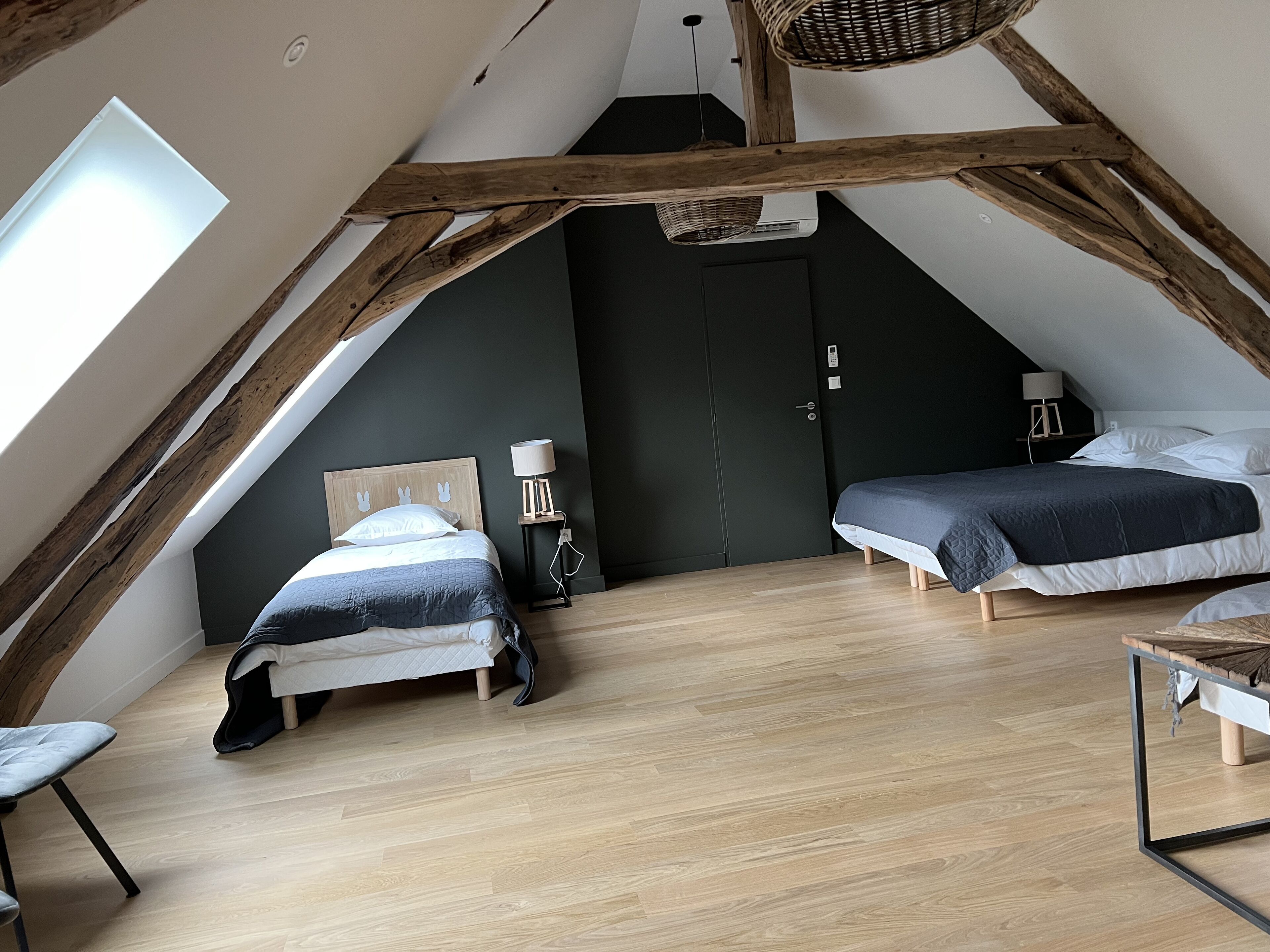 A bedroom with two beds, wooden beams, a skylight, a dark wall, and a small table with a lamp.