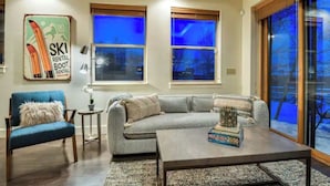 A cozy living room with a blue armchair, a gray sofa, and a coffee table with a board game. There are two windows with wooden blinds, one of which has a blue tint. A ski rental sign is visible on the wall. - skiNewpark33: Walkable w/View, Deck, Hot Tub, Garage (Park City)