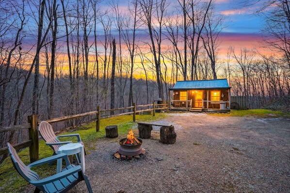 A cabin with a fire pit and Adirondack chairs in a forest setting.