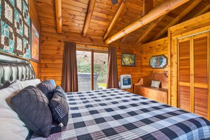 Een slaapkamer in een houten blokhut, met een bed, een houten hoofdbord, een gebogen sprei, een houten bankje en een raam met gordijnen.