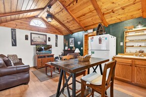 A cozy kitchen with wooden beams, a dining table, a refrigerator, and a shelf with items.