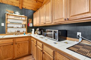 A kitchen with wooden cabinets, a white countertop, a microwave, a coffee maker, and a shelf with various items.