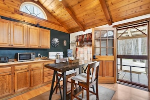 A wooden interior with a dining table, chairs, cabinets, a microwave, and a window with a view of trees.