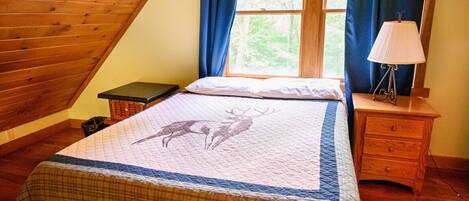 Une chambre avec un plafond en pente en bois, un lit orné d’un motif matelassé représentant un cerf, une lampe de chevet et une fenêtre avec des rideaux.