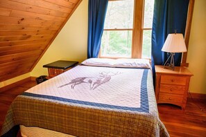 Quarto com teto de madeira inclinado, cama com estampa de veado acolchoado, luminária de cabeceira e janela com cortinas.