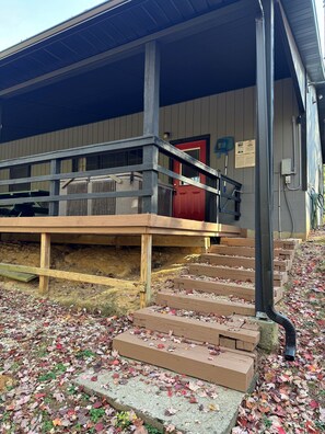 A building with a covered outdoor area, wooden steps leading up to a red door, and a sign on the wall. - Lazy Lane Cabins - Blue Jay (South Bloomingville)