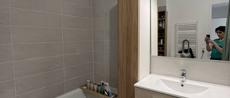 A modern bathroom with a bathtub, a vanity with a mirror, and a shelf with toiletries.