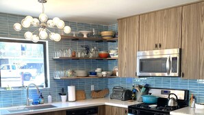 A modern kitchen with a central island, wooden cabinets, a stainless steel microwave, and a blue tiled backsplash. - Updated Mid-Century beach house --10 minute walk to the beach (Carlsbad)
