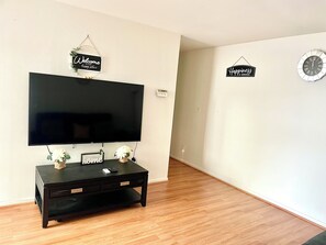 Una sala de estar con un televisor de pantalla plana sobre un mueble negro, piso de madera y dos adornos de pared con frases motivacionales.