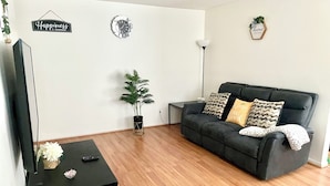 Un salon avec un canapé noir, un téléviseur posé sur un meuble noir, une plante en pot et deux éléments de décoration murale.