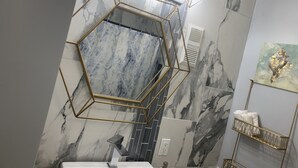A bathroom with a hexagonal mirror, marble walls, a gold-framed light fixture, and a towel rack.