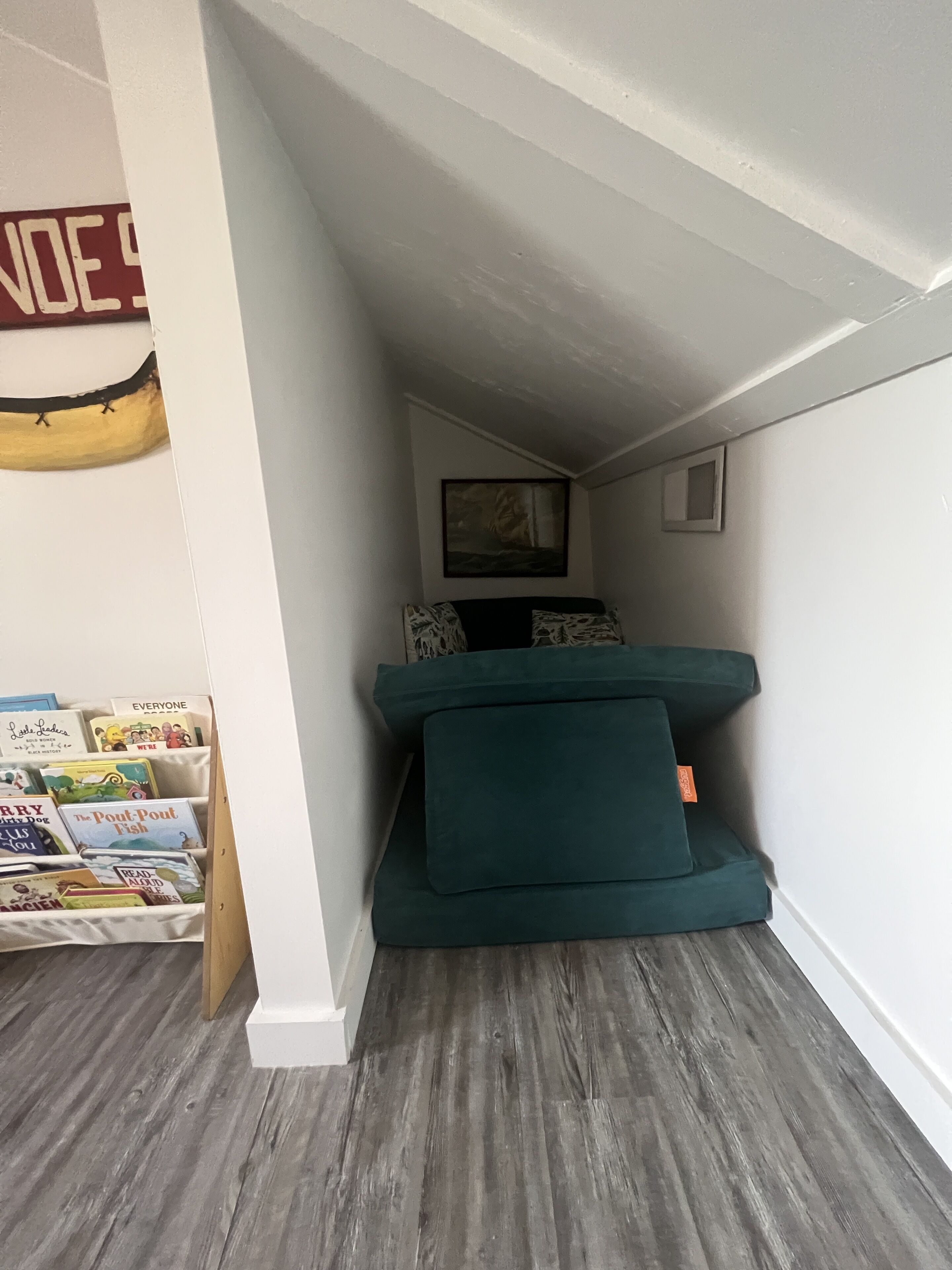 Een kleine kamer met een blauwgroene slaapbank, een klein lijstwerk met een afbeelding aan de muur, en een boekenplank vol boeken.
