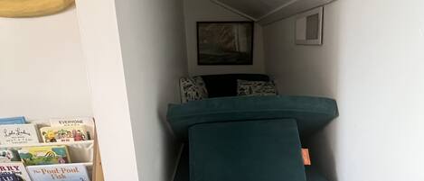 Een kleine kamer met een blauwgroene slaapbank, een klein lijstwerk met een afbeelding aan de muur, en een boekenplank vol boeken.