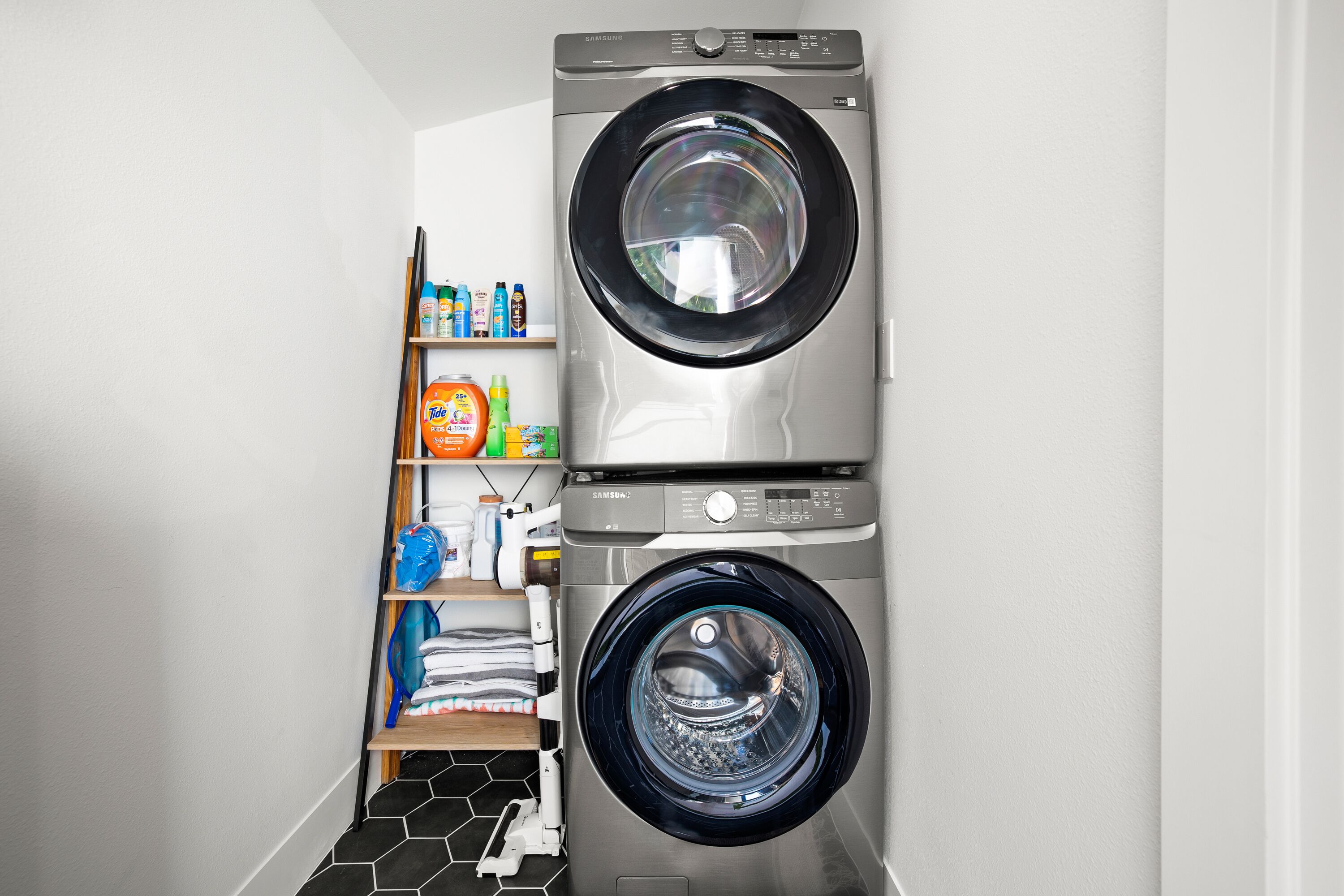 A laundry room with two stacked front-loading washing machines and a shelf with various cleaning products.