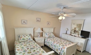 Een slaapkamer met twee bedden, een plafondventilator, een dressoir, een televisie en ingelijste foto's aan de muur.
