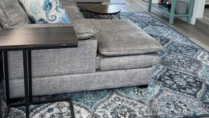 A living room with a sectional sofa, a small coffee table, a patterned rug, a TV mounted on the wall, and a side table with decorative items. - Newly Renovated 1 Bed/1.5 Bath Mobile Home Near Ft. Myers Beach, Florida (Fort Myers)