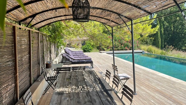 Uma área externa coberta com piscina, deck de madeira, espreguiçadeiras e uma mesa com bancos.