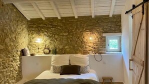 Quarto com parede de pedra, cama com roupa de cama branca, luminária pendente e teto de madeira.