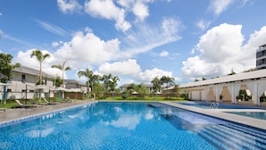 A swimming pool with lounge chairs and umbrellas, surrounded by palm trees and buildings.