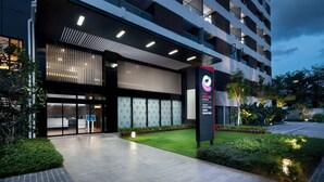 A modern hotel entrance with a glass facade, outdoor seating area, and a well-lit pathway.
