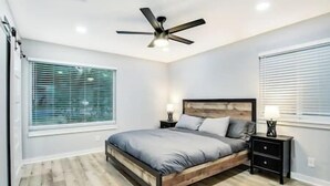 Une chambre à coucher avec un lit, un ventilateur de plafond, une table de chevet et une fenêtre avec des stores.
