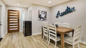 Un coin repas avec une table en bois et des chaises blanches, une cheminée et un mur sur lequel est inscrit le mot « Another » en lettres bleues.