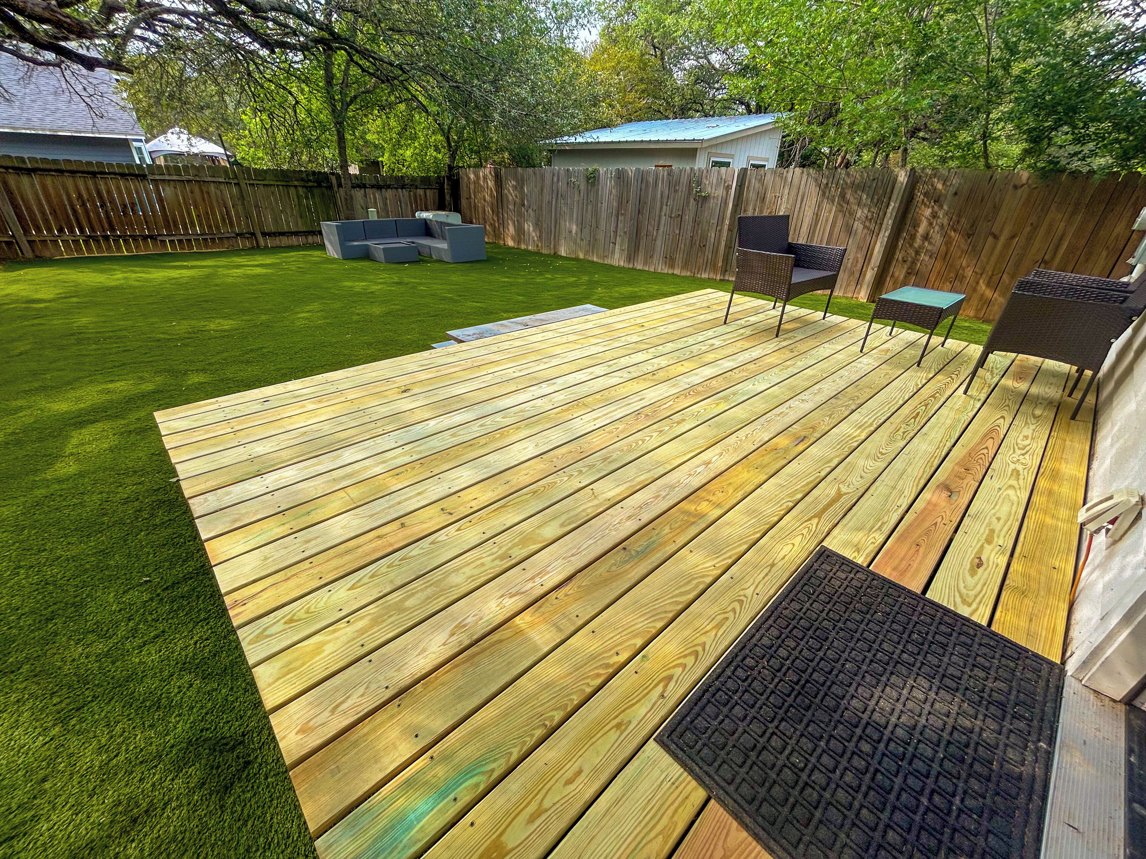 Una terraza de madera con jardín césped, valla de madera y mobiliario exterior.