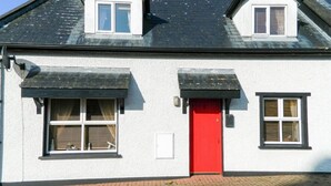 A white building with a red door, two windows with shutters, and a small dormer window. - Charming cottage in a traditional Irish fishing village. (Greencastle)