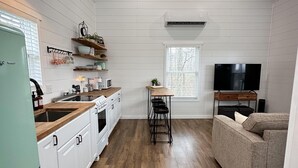 A compact kitchen with white cabinets, a green refrigerator, wooden countertops, a dining table with stools, a TV, and a window with blinds.