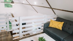 A loft-style room with a white ceiling, wooden beams, a gray sofa with a yellow pillow, and a white coffee table with a plant.