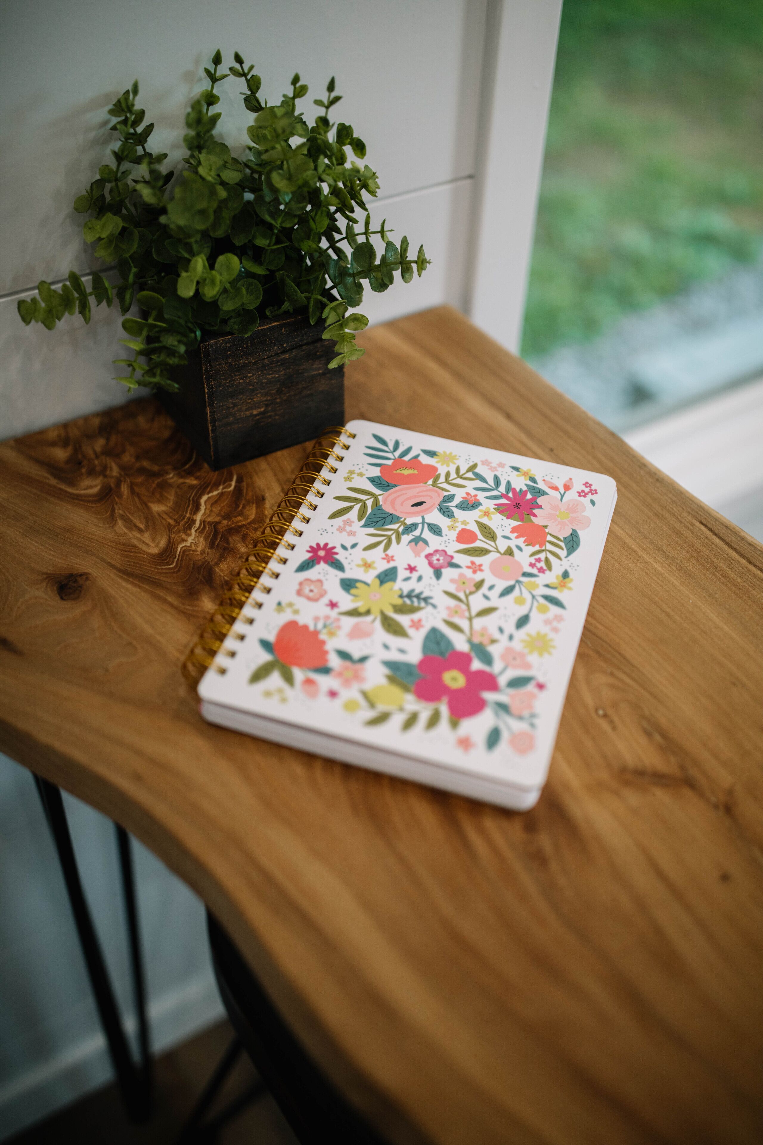 Um caderno com capa floral sobre uma mesa de madeira, ao lado de um vaso com planta.