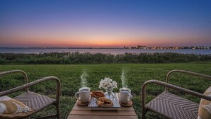 Auringonlaskunäkymä vehreältä terassilta, jossa on kaksi rottingista tuolia, tarjotinalusta leivonnaisineen ja kaksi kahvikuppia.
