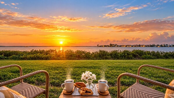 A sunset view with a table set for two, including a cup of coffee, a pastry, and a vase of flowers.