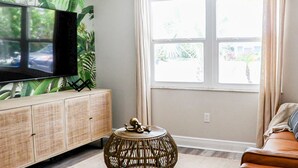 A living room with a flat-screen TV mounted on a bamboo-patterned cabinet, a wicker side table, and a view of a street through a large window. - True Tropical Escape at The Coconut House (Fort Pierce)