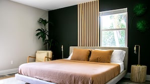 A modern bedroom with a large bed, a chair, a potted plant, and a window with blinds. - True Tropical Escape at The Coconut House (Fort Pierce)
