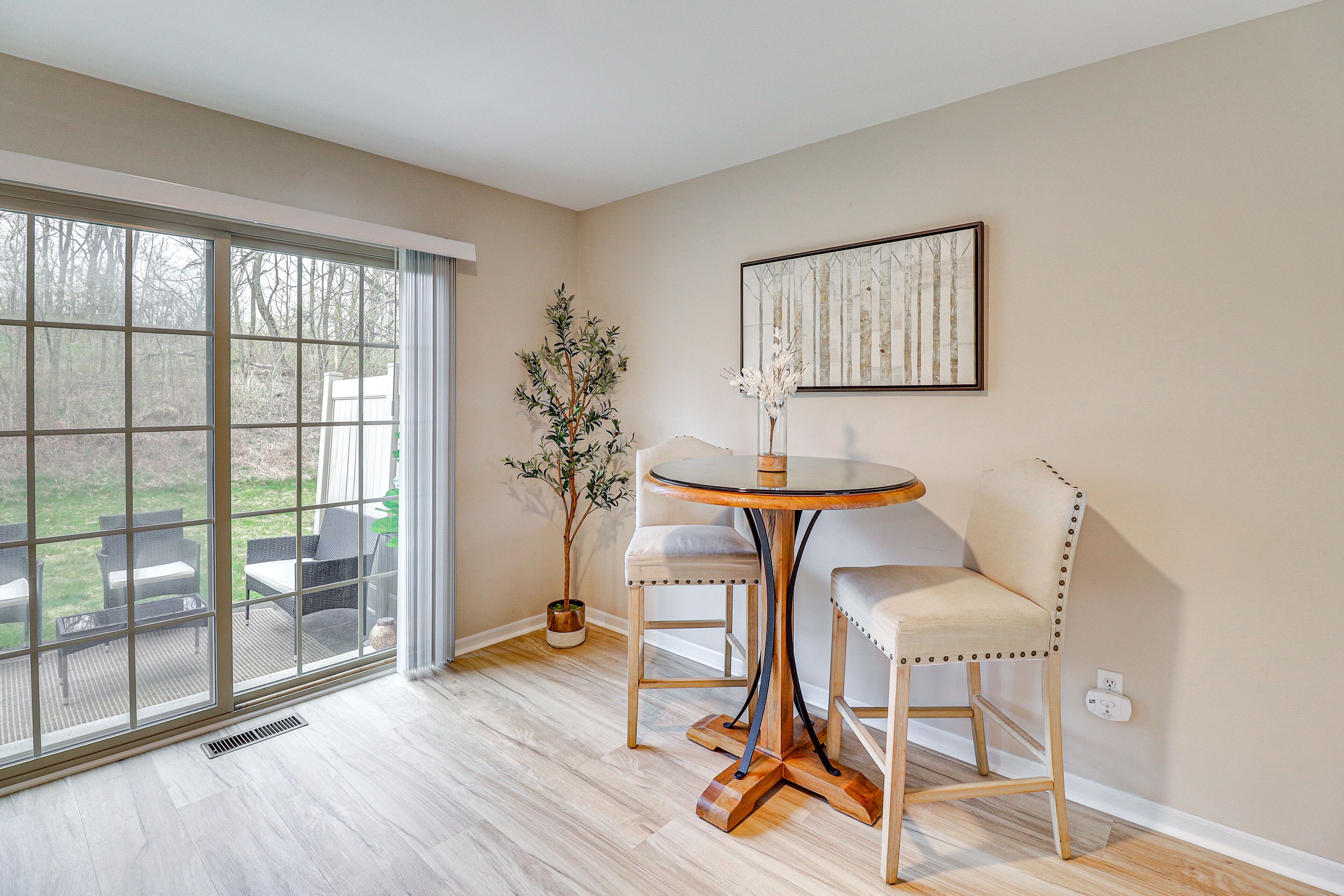 Ein Zimmer mit einer großen Glasschiebetür, die auf eine Terrasse führt, einem runden Tisch mit zwei Stühlen, einem Blumentopf und einem gerahmten Bild an der Wand.