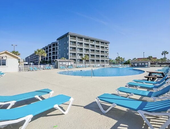 A pool area with blue lounge chairs, a large building in the background, and a clear sky. - NEW! Ocean-Front Resort, Steps From Beach, 6 pools and Lazy River (Myrtle Beach)