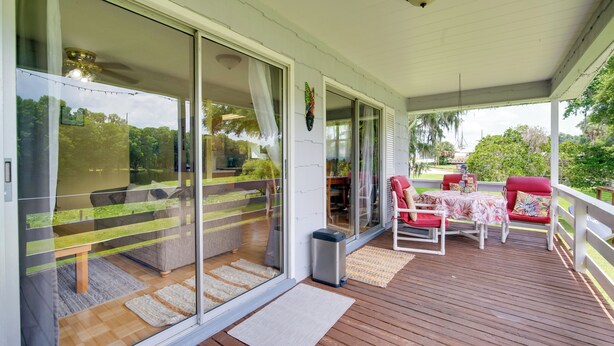 Área externa coberta com deck de madeira, portas de correr de vidro que dão para um espaço interno com sofá e mesa de jantar.
